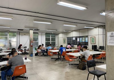 Salão BEE - Biblioteca da Escola de Engenharia e do Instituto de Computação. Na foto, salão com usuários estudando, sentados à mesas de tampo branco e cadeiras na cor laranja. Ao fundo a direita computadores sendo utilizados pelos estudantes.